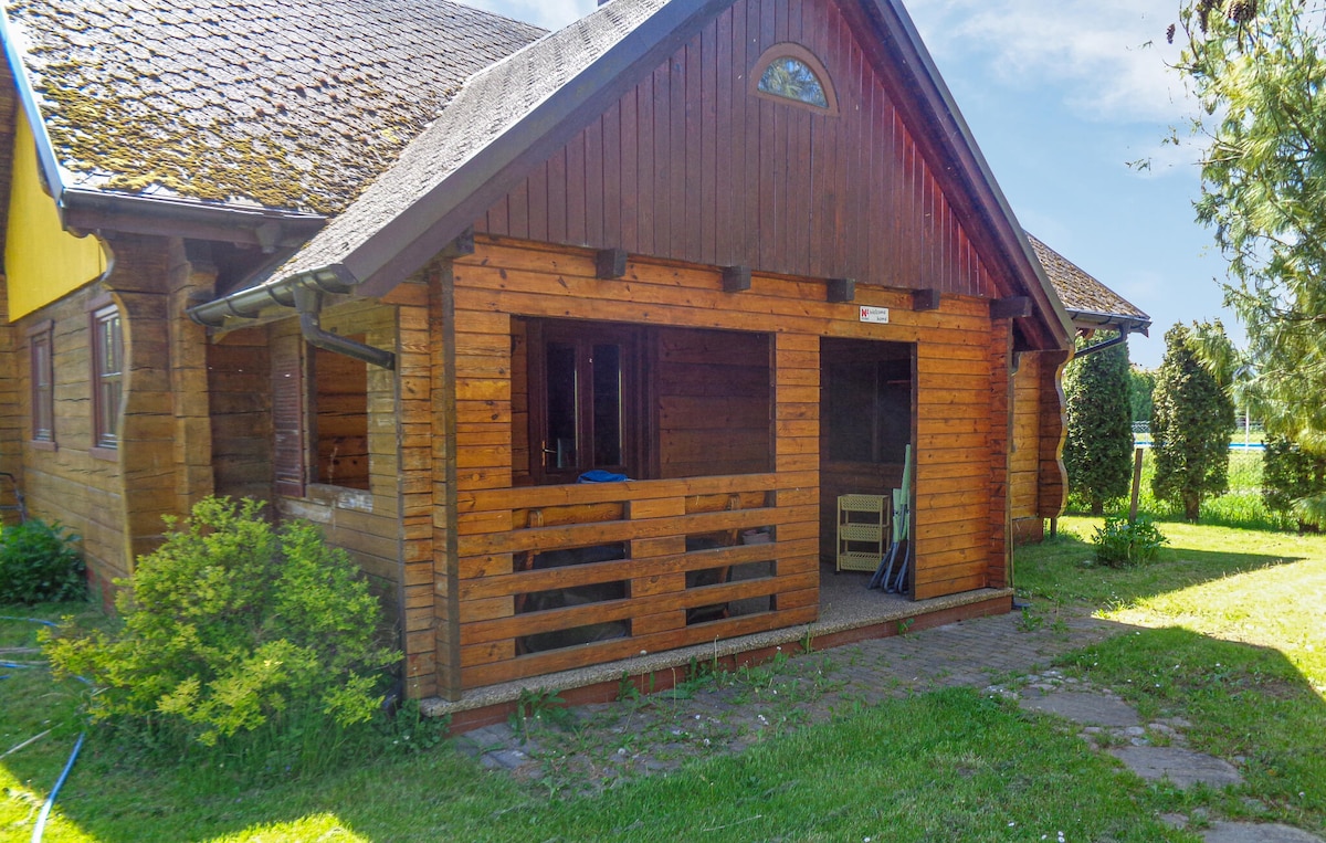The exterior of the wooden beam house is shown, featuring a covered porch with a wooden railing. The surrounding grassy area includes shrubs and trees, providing a natural setting. Sunlight brightens the scene, highlighting the rustic charm of the structure.