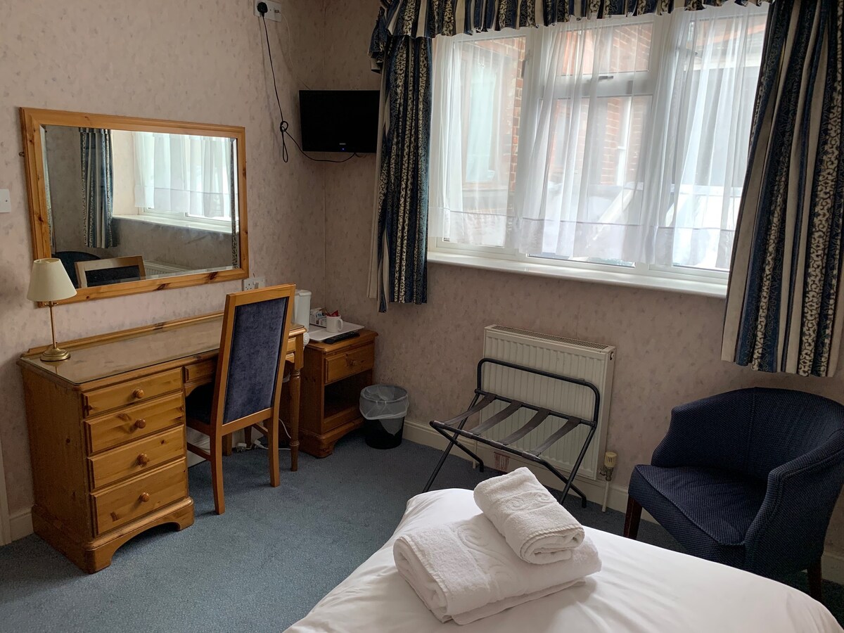 A bright guest room features a spacious bed with fresh linens. A wooden vanity with a mirror and a chair is positioned against the wall. An armchair, a folding luggage rack, and a flat-screen TV are also present, while natural light enters through the sheer-curtained window.