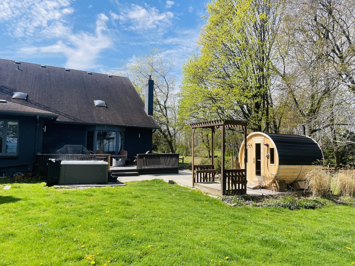 The exterior features a spacious backyard with a wooden deck and lush green grass. A traditional wood sauna is placed beside a seating area, complemented by trees and clear blue skies in the background. Sunlight brightens the scene, creating a serene outdoor setting.