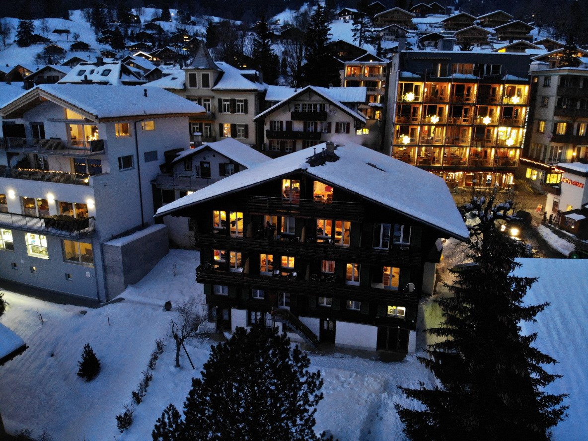 A snow-covered landscape reveals the hotel building, characterized by traditional wooden architecture. Surrounding structures are visible, showcasing multi-story designs with illuminated windows. Snow blankets the ground, enhancing the serene winter atmosphere.