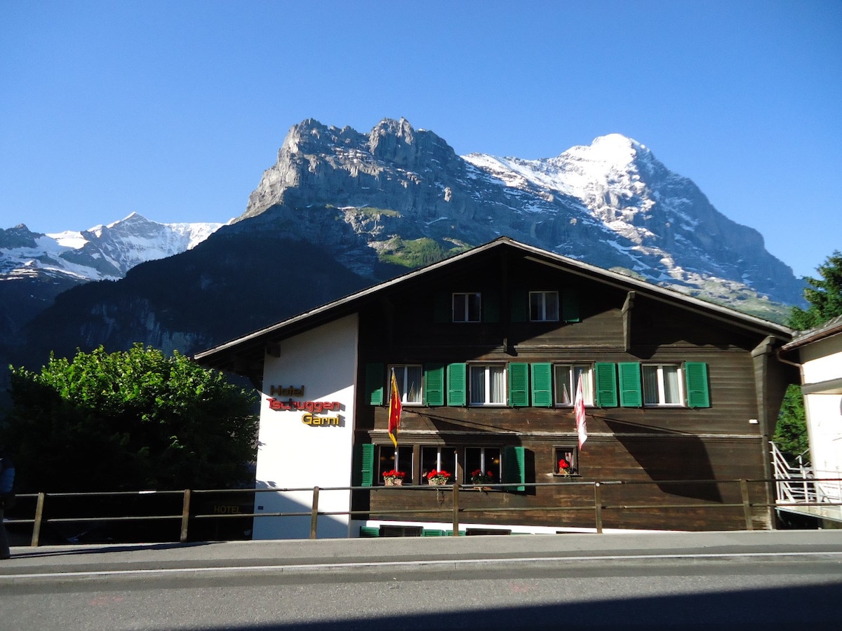 A wooden hotel building with green shutters is positioned in front of a stunning mountain backdrop. Brightly colored flower boxes adorn the windows, and flags are displayed outside. The surrounding area features lush greenery, and the sky is clear with soft sunlight illuminating the scene.