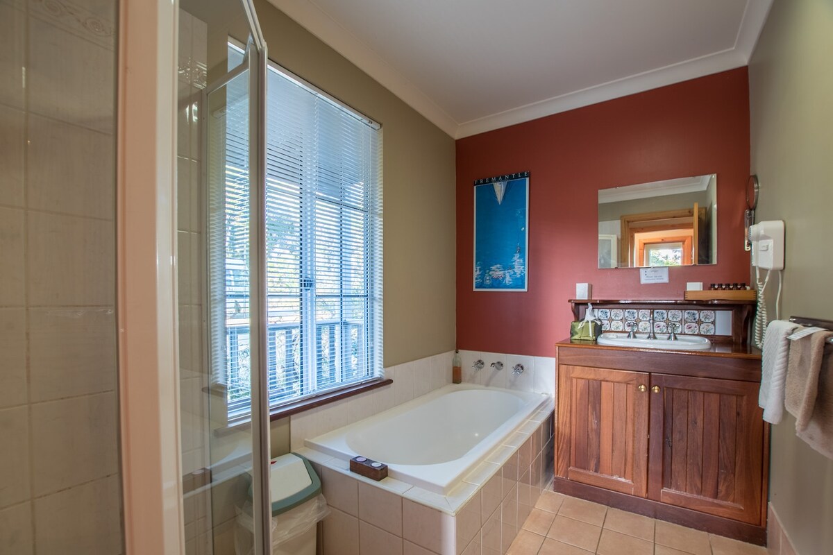 The bathroom features a spacious bath adjacent to a walk-in shower, with natural light streaming through a window covered by horizontal blinds. A wooden vanity with a mirror adds warmth, while tasteful wall colors create a calming ambiance.