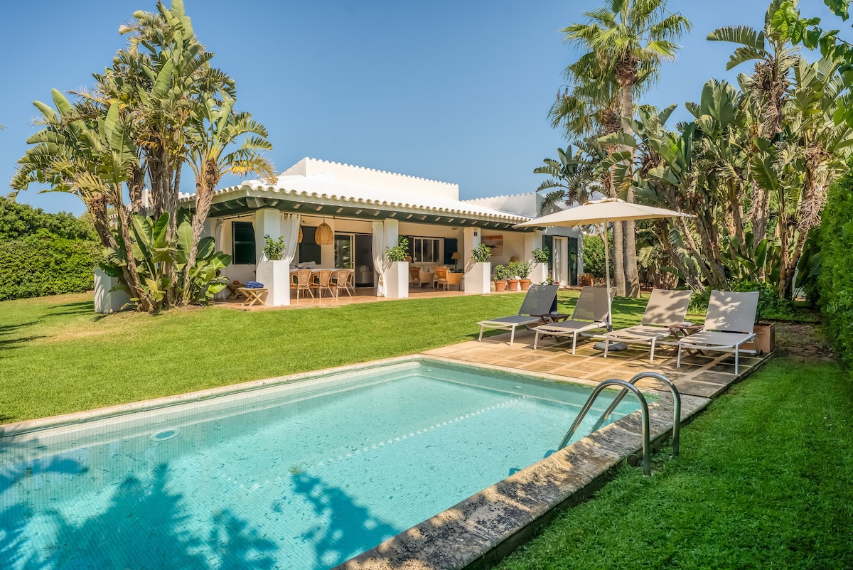 A private outdoor area features a swimming pool surrounded by sun loungers. Lush greenery and palm trees create a serene environment. The charming chalet is visible in the background, complemented by a covered terrace and dining space. A bright blue sky completes the inviting scene.