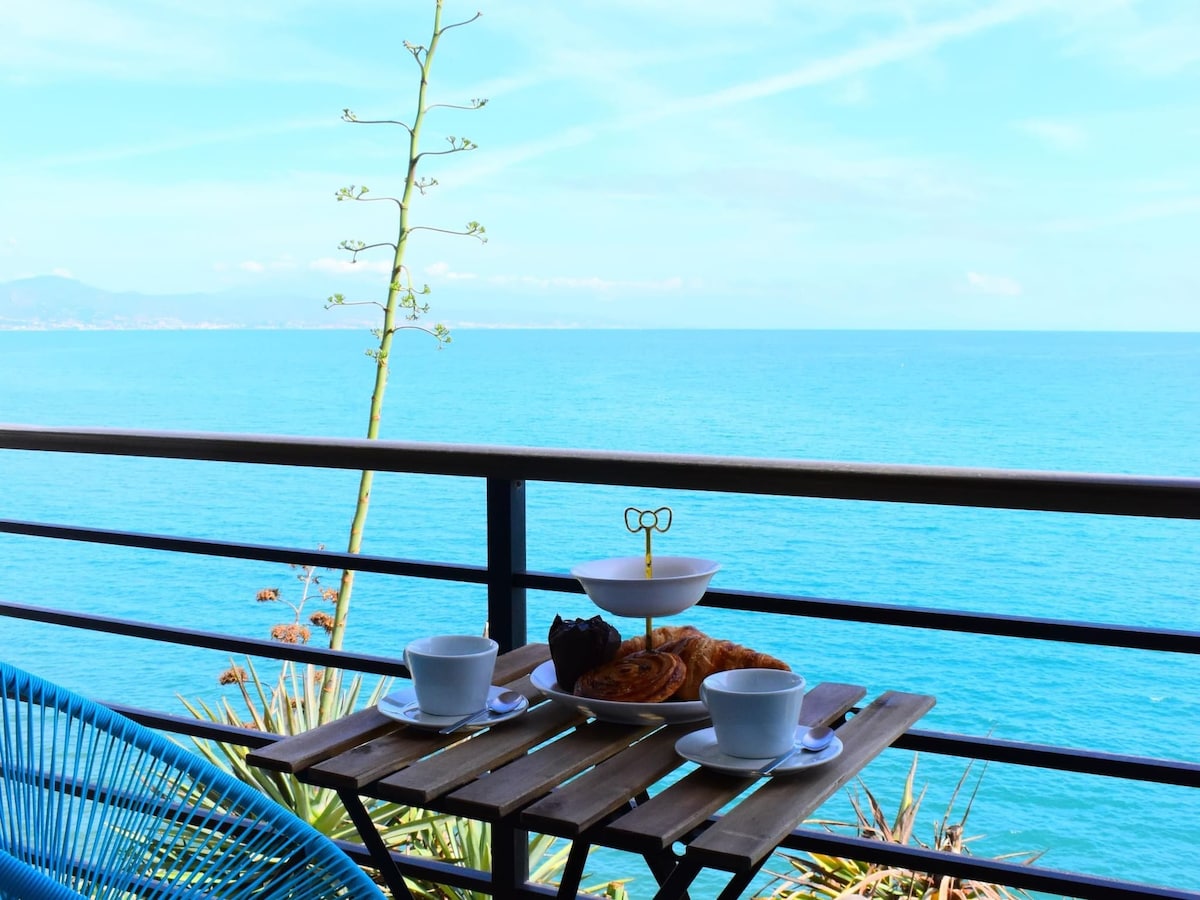 A small wooden table is set on a balcony with a clear view of the ocean. Two cups and a bowl of pastries are arranged on the table, framed by vibrant greenery. The sea stretches into the horizon under a bright blue sky.