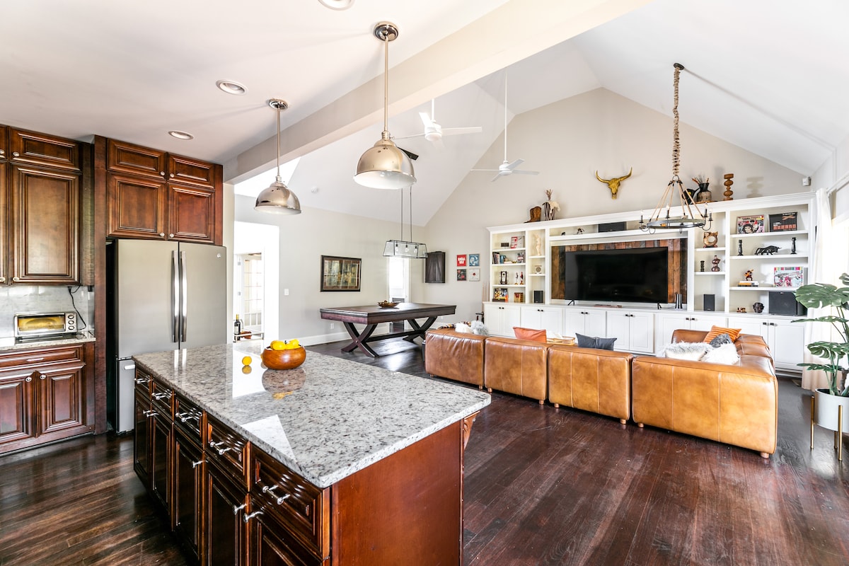 A spacious living area is highlighted by a high, vaulted ceiling and polished wooden floors. A large kitchen island with granite countertops features a fruit bowl. Comfortable leather sectional seating is positioned in front of a wall-mounted television and built-in shelving.