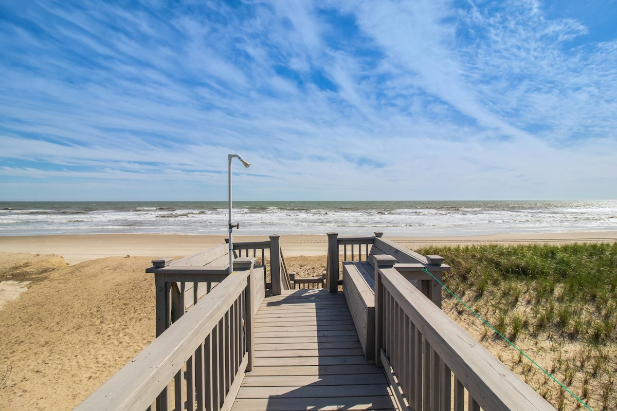 A wooden walkway leads from the property to the sandy beach, providing direct access to the ocean. An outdoor shower is visible, designed for rinsing off after beach visits. The scene highlights a clear blue sky with gentle clouds above the sparkling water.