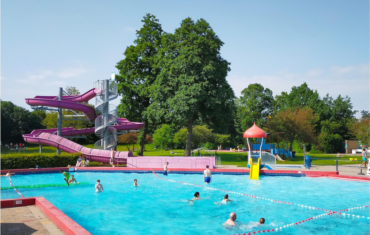 A recreational area features a large pool with swimmers enjoying the water. A vibrant water slide stands nearby, accompanied by a playful water play structure. Lush trees surround the space, providing shade and enhancing the outdoor atmosphere.