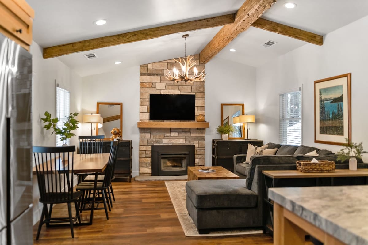 A modern living area features a stone fireplace beneath a wooden beam ceiling. A large television is mounted above the fireplace. Comfortable seating is arranged around a coffee table, and a dining space with black chairs is visible. Natural light enters through multiple windows.