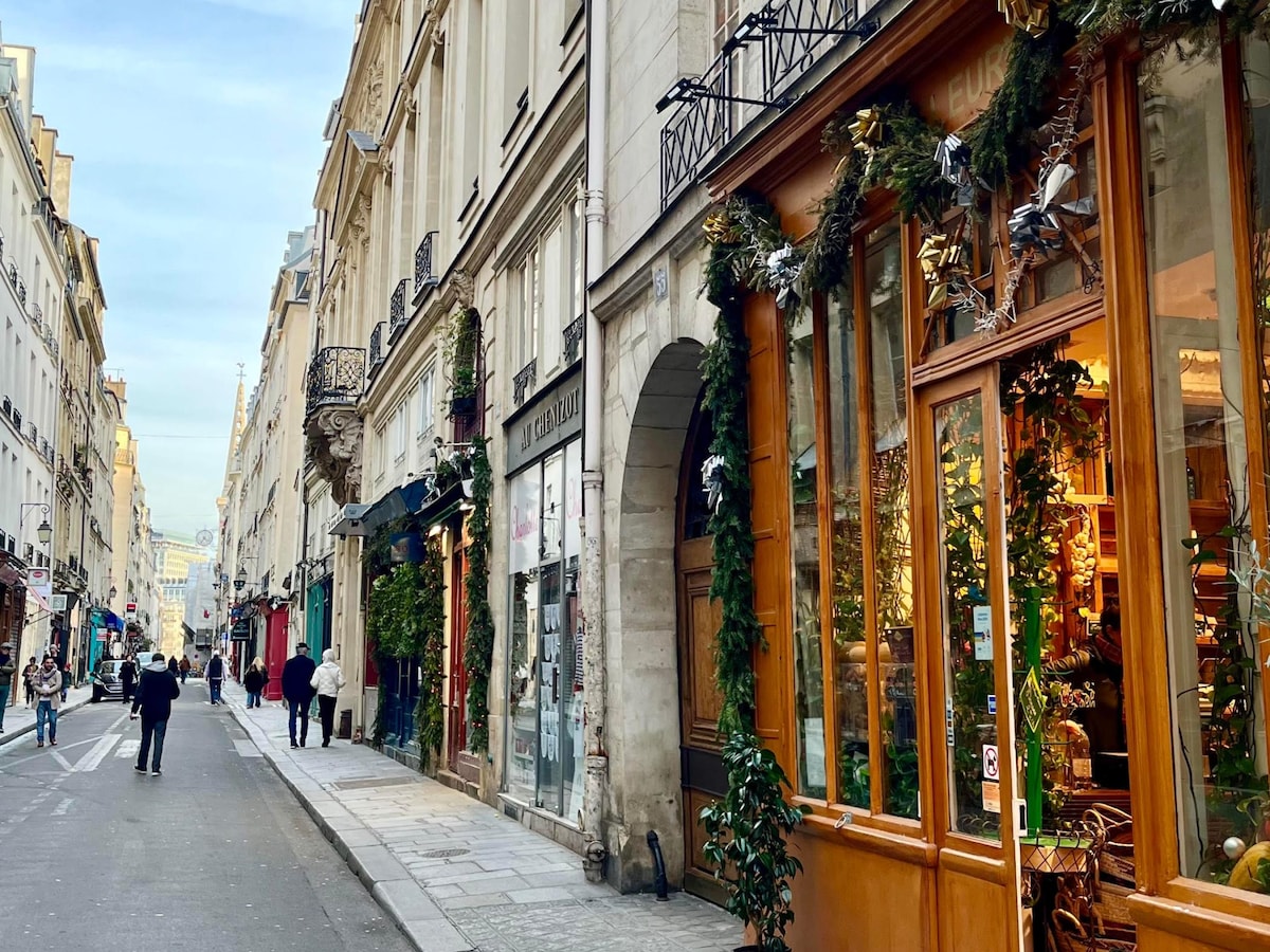 A charming street scene in Paris is captured, showcasing historic buildings adorned with festive greenery. The wooden-framed shop window displays inviting merchandise under warm lighting. Pedestrians are seen strolling leisurely, contributing to the vibrant atmosphere of the area.