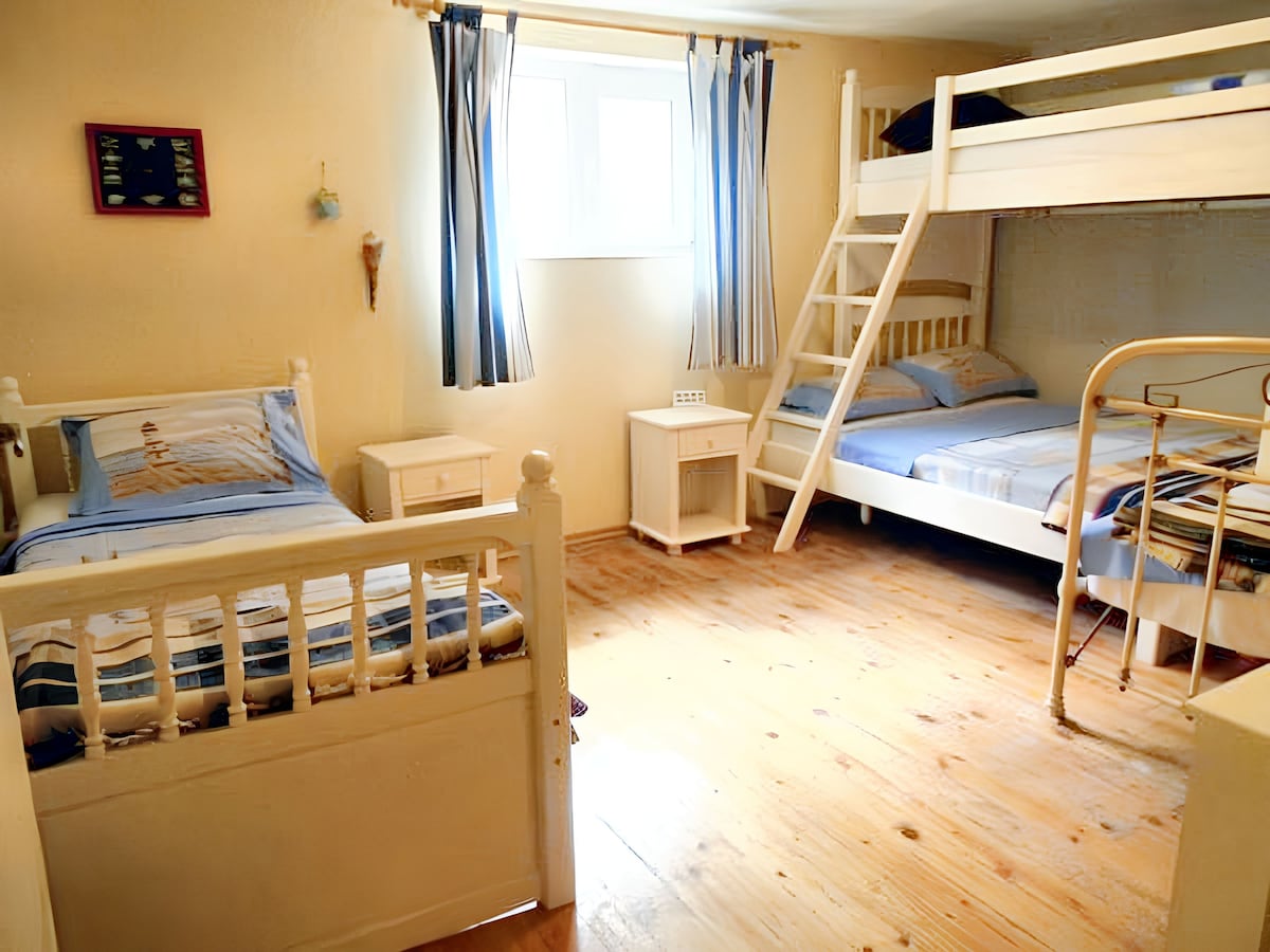 A room features a combination of beds, including a bunk bed and a single bed. Simple wooden furnishings in a light color complement the neutral walls and wooden floor. Natural light filters through a window, adorned with blue and white striped curtains.