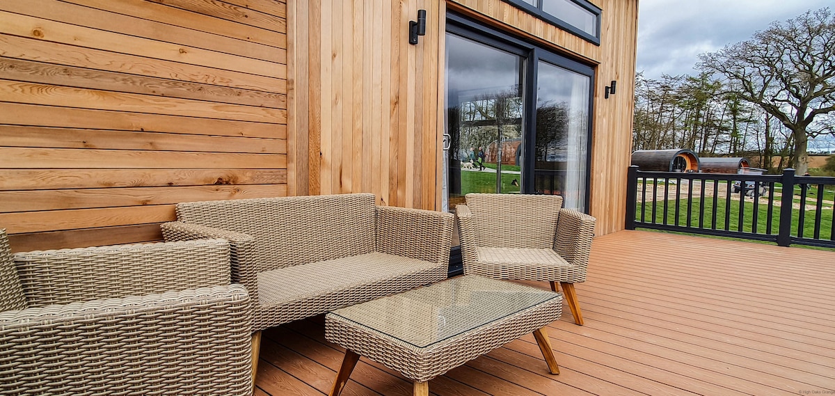 An outdoor seating area is presented with a rattan-style furniture set, including two armchairs and a small coffee table. The wooden decking and surrounding natural scenery create a relaxing environment, enhanced by large glass doors leading to the lodge's interior.
