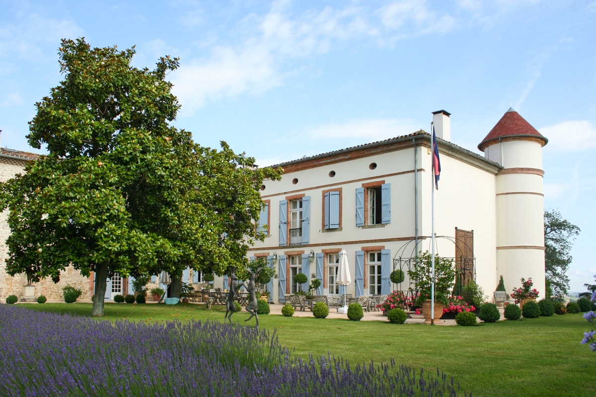 The château's exterior features a charming design with a turret and inviting entry. Lush gardens, adorned with flowering plants and manicured hedges, surround the property. A prominent tree offers shade, while lavender blooms create a vivid contrast in the foreground, enhancing the picturesque setting.