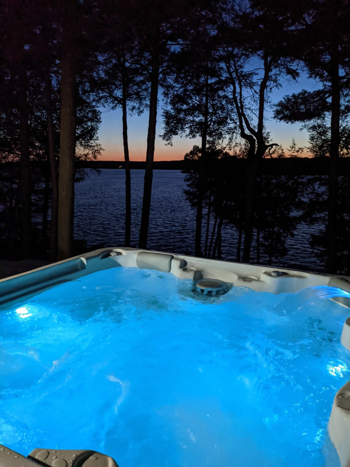 A hot tub is positioned on a patio overlooking a serene lake, illuminated with soft blue light. Trees frame the view, creating a private atmosphere during twilight hours, where the sky displays a gradient of colors as the sun sets beyond the water.