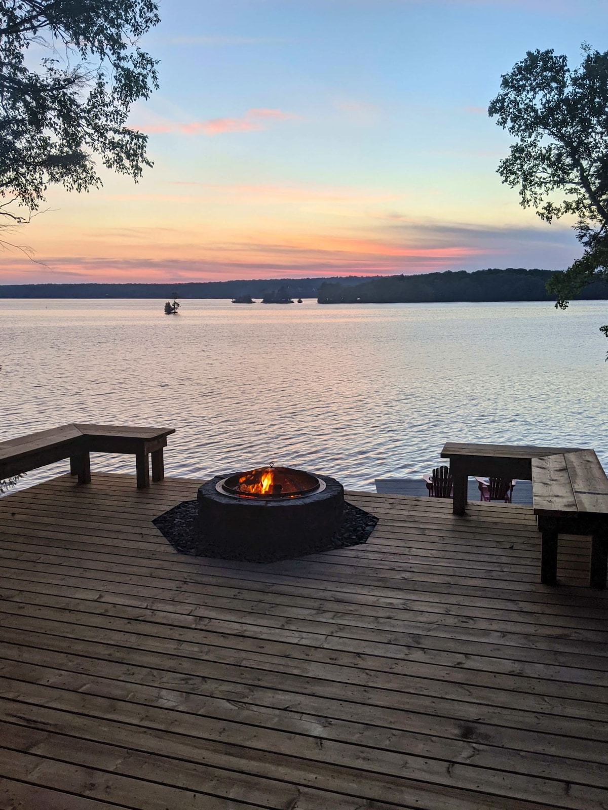 A wooden deck features seating arrangements around a centrally placed fire pit. The area is surrounded by trees, with a scenic view of the lake in the background, reflecting soft hues of sunset. Adirondack chairs are positioned nearby, inviting relaxation by the water.