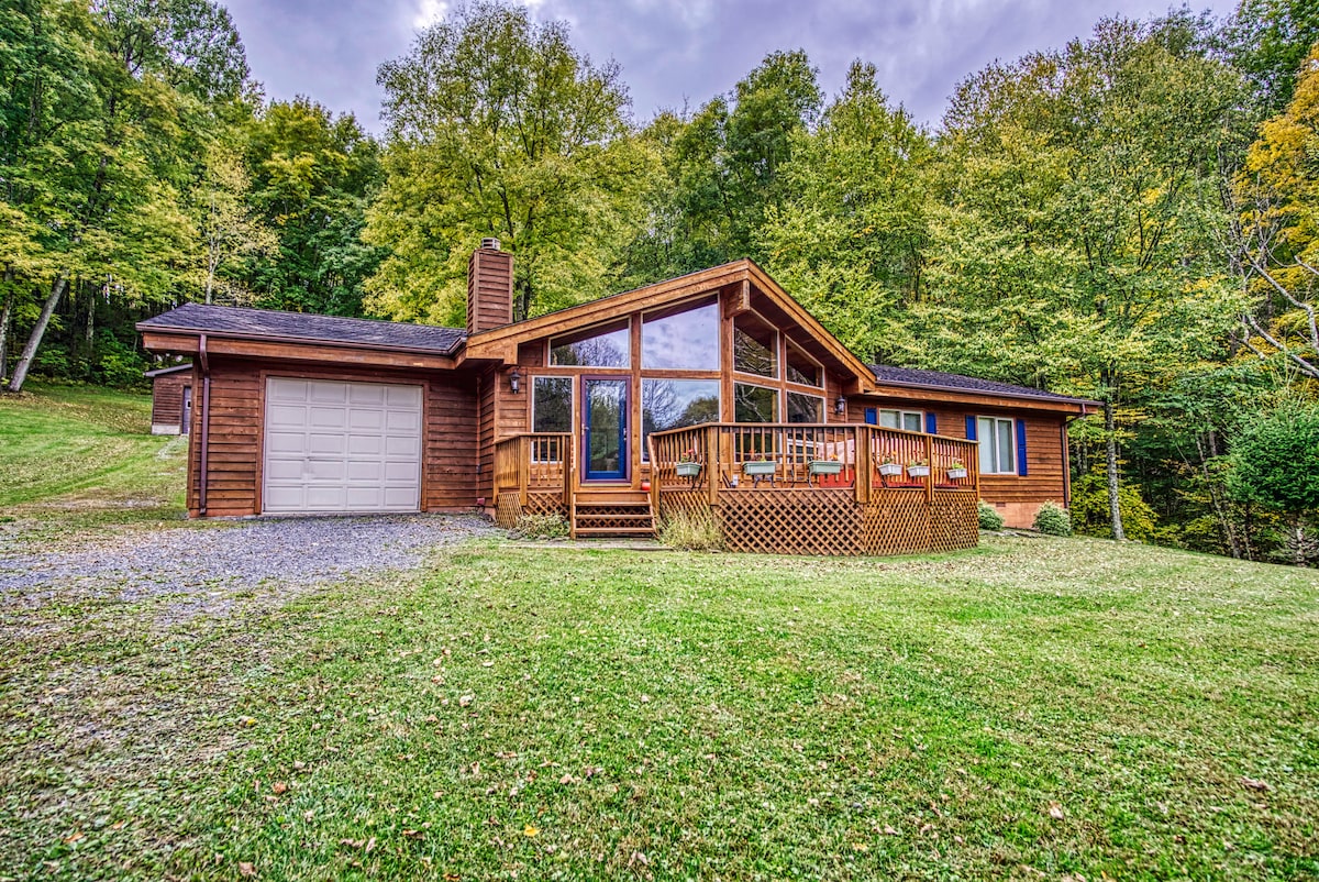 The wooden chalet is nestled among lush greenery, featuring a large front deck and expansive windows that offer views of the surrounding forest. A gravel drive leads to the attached garage, emphasizing the home's inviting nature and serene setting.