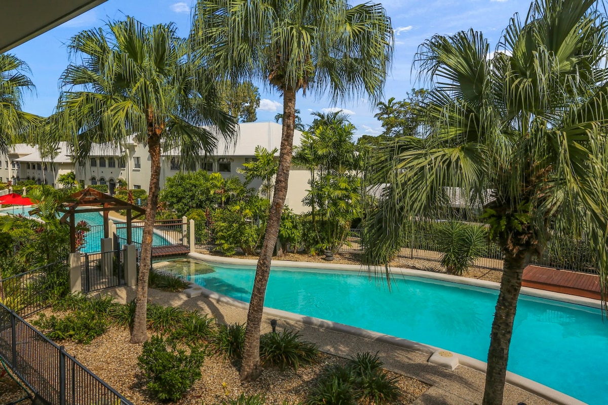 A serene view of the resort is captured, featuring a meandering swimming pool surrounded by tropical palm trees and lush greenery. The pool area is complemented by sun loungers, creating a relaxing atmosphere. Bright blue skies and the distant edges of the resort buildings are also visible.