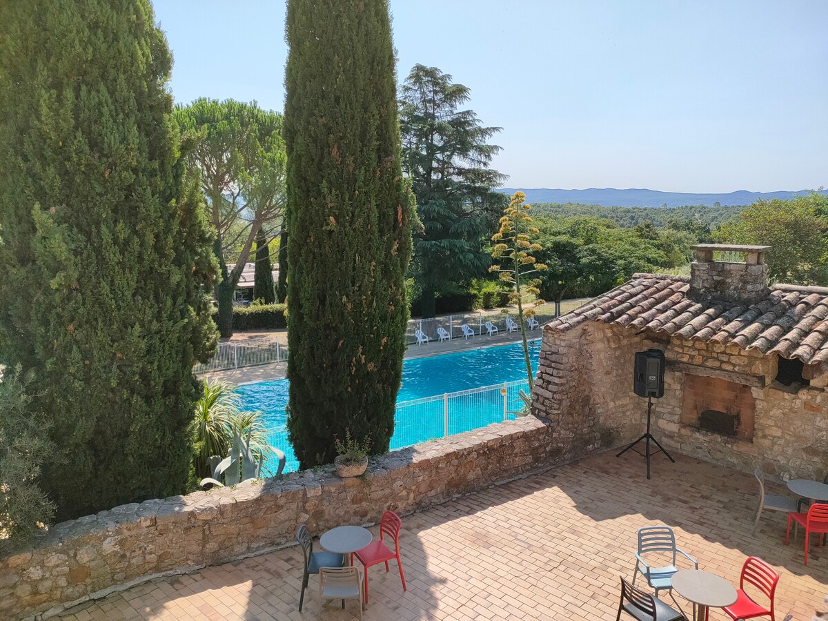 The outdoor area features a tranquil stone terrace with several small tables and colorful chairs. The view includes a refreshing swimming pool surrounded by tall trees, complemented by scenic landscape beyond.