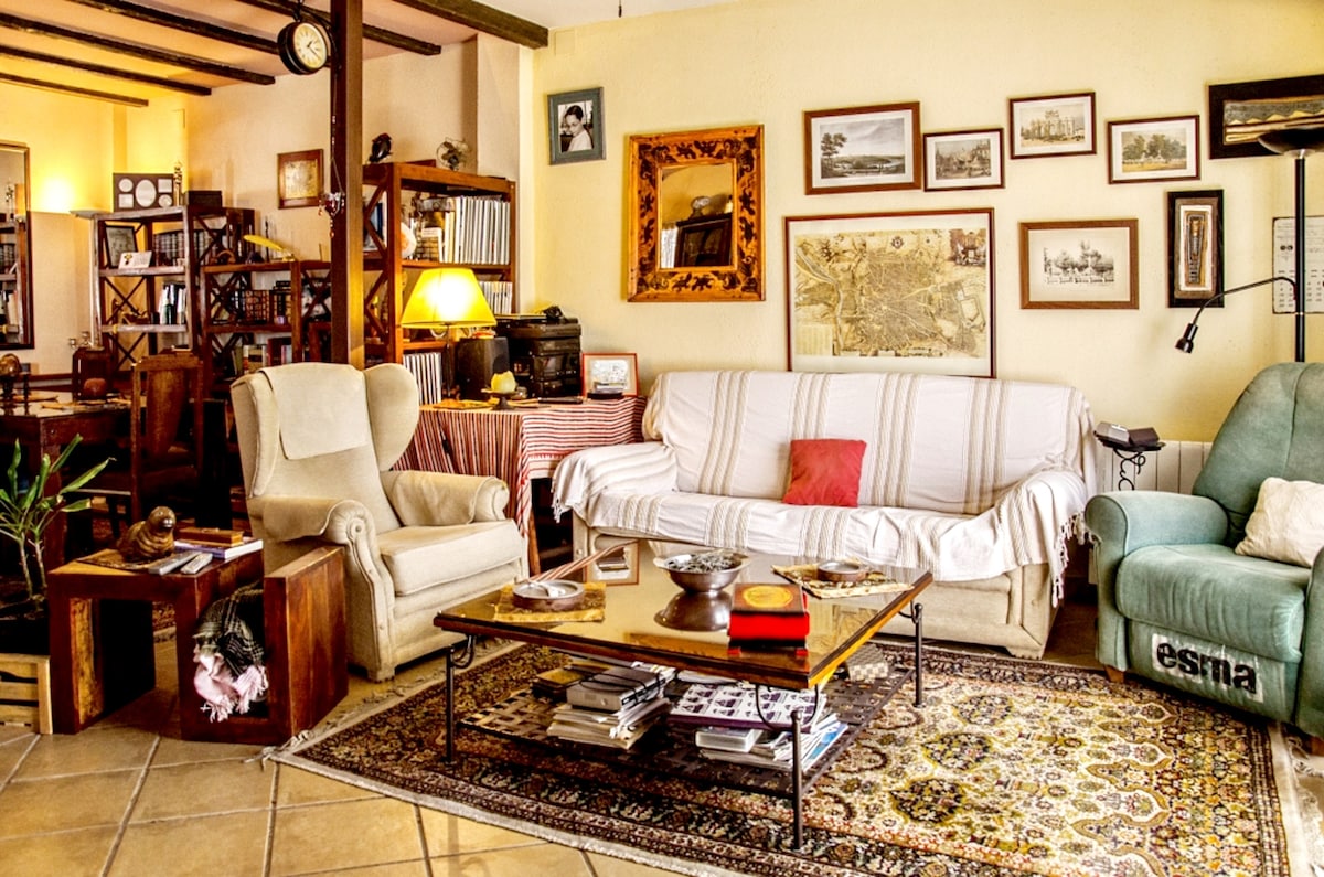 A spacious living area is presented with a comfortable seating arrangement featuring a light-colored sofa, armchairs, and a coffee table. Surrounding the space are shelves filled with books, along with various framed pictures enhancing the homey atmosphere. Natural light filters through the windows.