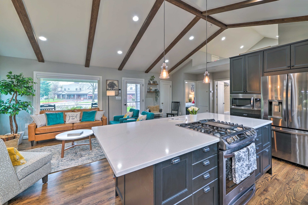 A modern kitchen features a spacious island with a gas range and stainless steel appliances. Exposed wooden beams soar above the open living area, which is furnished with a cozy sofa, accent chairs, and a large window offering views of the outdoor space.