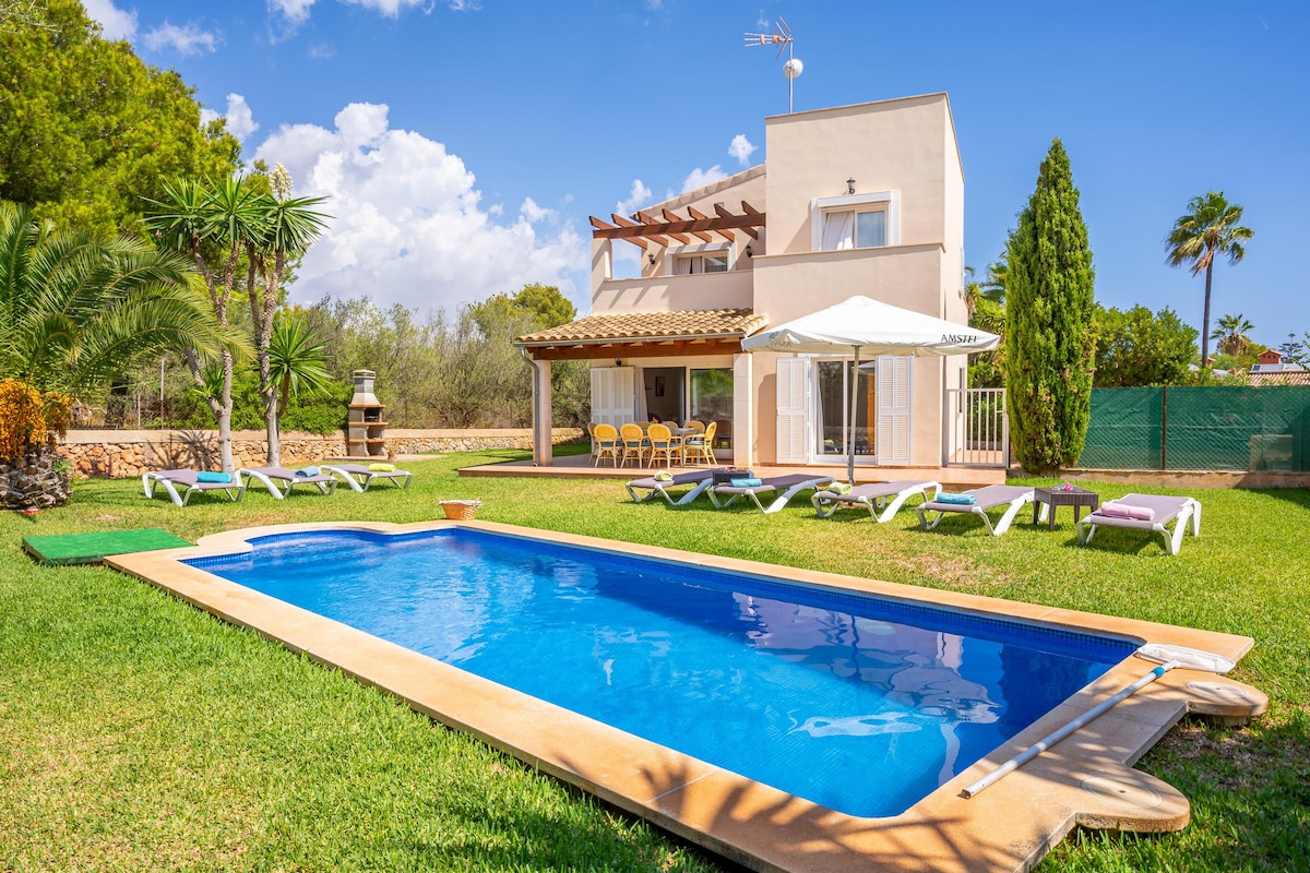 A private outdoor area is displayed, featuring a blue swimming pool surrounded by manicured grass. Lounge chairs are arranged around the pool. A two-story holiday home sits in the background, with a covered terrace and a parasol for shade. Lush greenery enhances the setting.