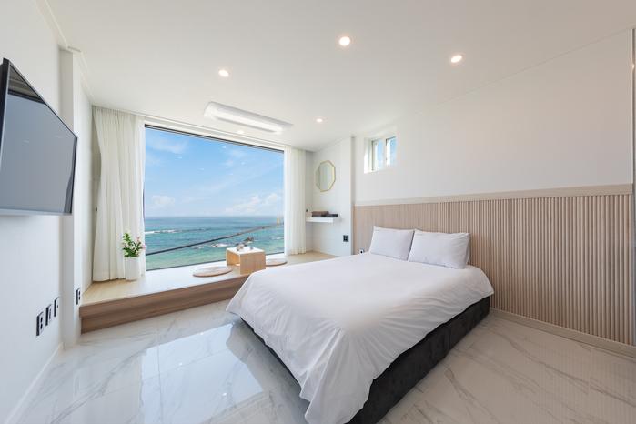 A bright sleeping area features a spacious bed with crisp white linens, positioned against a backdrop of large windows offering ocean views. Natural light fills the room, which is characterized by minimalist décor and soft neutral tones, complemented by a sleek wall-mounted television.