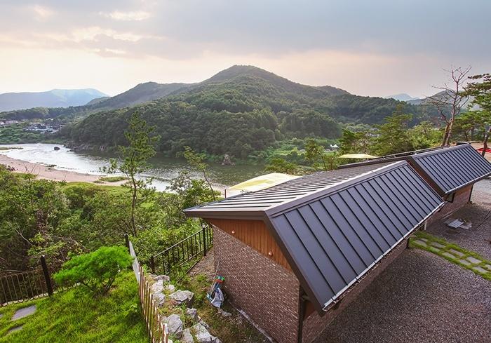 A separate accommodation showcasing a sloped roof and stone pathway is positioned amid lush greenery. The surrounding landscape features rolling hills and a river, creating a serene backdrop. The scene is complemented by soft hues of the setting sun filtering through the clouds.