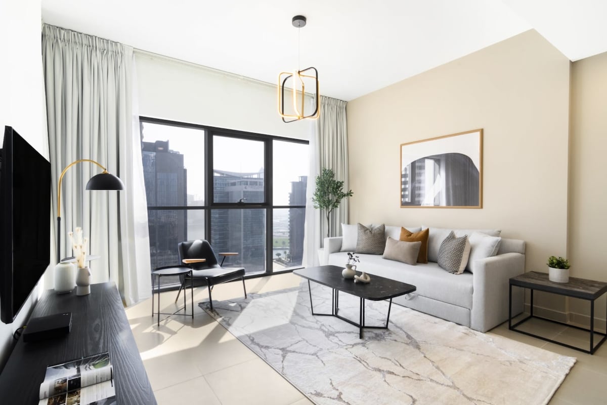The living room is designed with minimalist modern aesthetics, showcasing a comfortable sofa and an armchair. A coffee table sits central on a light area rug. Large windows allow natural light to penetrate, providing a view of the urban surroundings. Decorative plants add a touch of greenery.