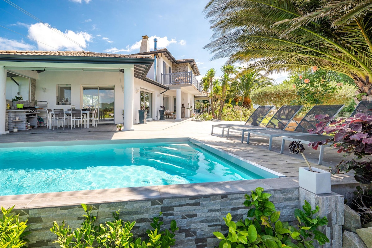 An inviting outdoor area features a private swimming pool surrounded by a sun deck. Loungers are positioned for relaxation, while a terrace overlooks landscaped gardens. The villa's exterior reflects a harmonious blend of modern design and natural elements, complemented by lush greenery and palm trees.