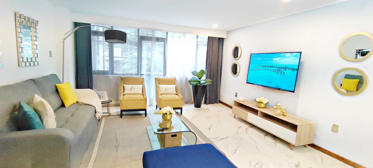 A modern living room features a gray sofa accompanied by two yellow armchairs. A flat-screen TV is mounted on the wall, while natural light filters through large windows covered with light curtains. A coffee table holds decorative items, complementing the contemporary decor.
