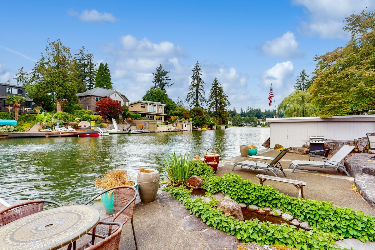 The serene riverfront view features a peaceful waterway lined with trees and residential homes. A furnished patio includes a round table and chairs alongside lounge chairs. Lush greenery surrounds the area, creating a tranquil outdoor space perfect for relaxation by the water.