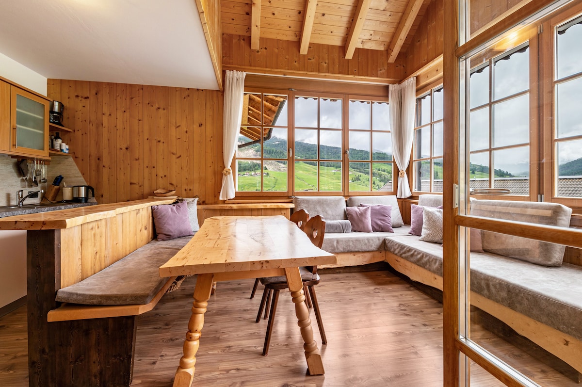 The living area showcases a wooden dining table surrounded by comfortable seating. Large windows frame scenic views of the landscape, while soft cushions complement the seating area. The kitchen features wooden cabinetry, blending rustic charm with modern functionality.