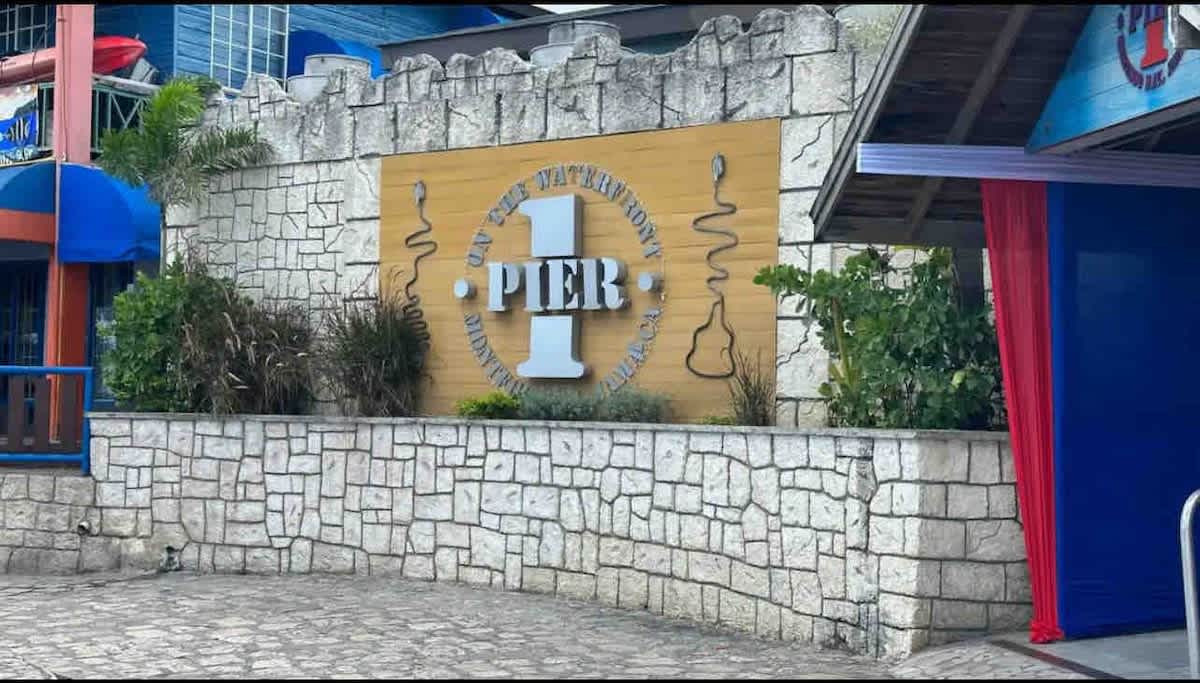 The entrance to a lively venue features a stone wall prominently displaying the number '1' in a bold font, accompanied by the word 'PIER.' Surrounding greenery adds a touch of nature, while the building's colorful awnings suggest a vibrant atmosphere.