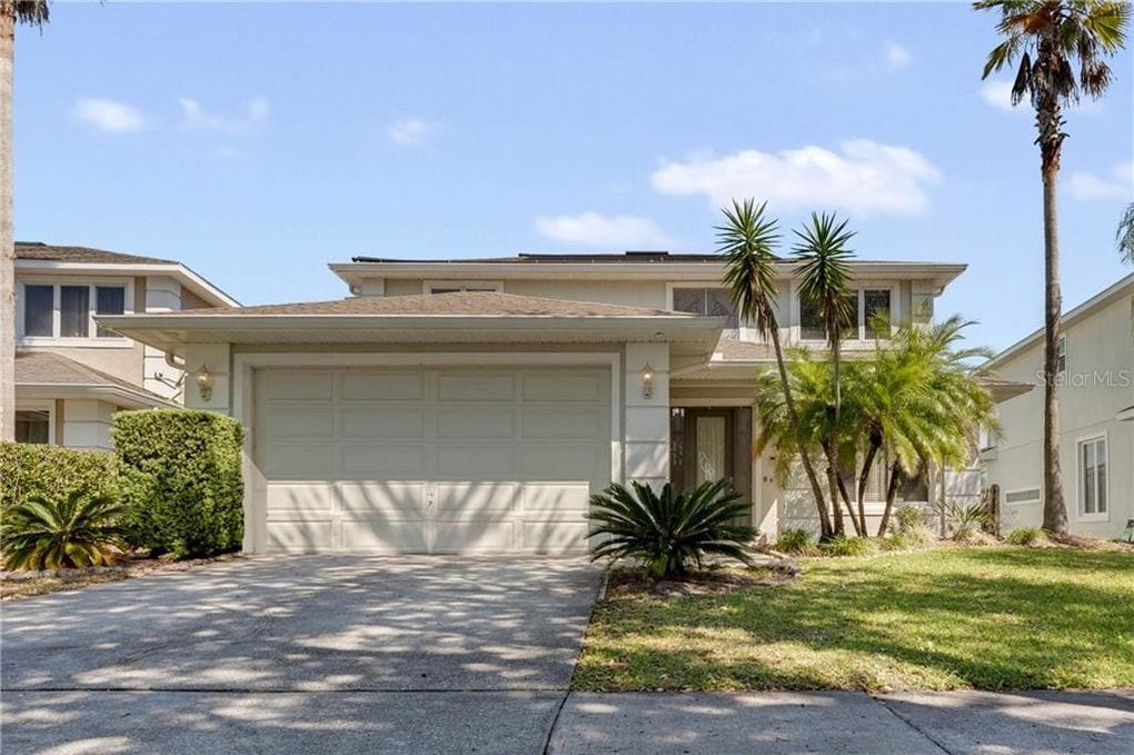 The exterior of the home features a two-story design with a wide driveway and a garage. Lush greenery, including palm trees and shrubs, flanks the entrance, enhancing the welcoming appearance. A clear sky provides ample natural light, highlighting the homeās muted facade.