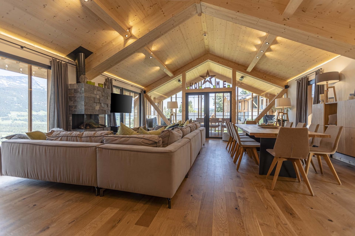 A spacious living area features a modern design with large windows offering a panoramic view of the mountains. A comfortable sectional sofa is arranged around a stone fireplace, while a wooden dining table is set with stylish chairs. Natural light fills the room, highlighting the wooden ceiling.