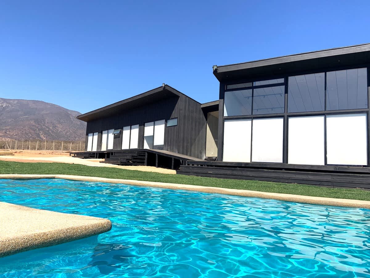 A modern minimalist house is depicted beside a clear blue pool. Floor-to-ceiling windows reflect the open sky and surrounding mountains. A spacious deck leads to the grassy area, creating a seamless connection between the home and outdoor space.