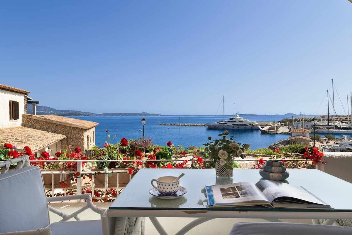 A covered terrace is adorned with flowering plants and a table set with a cup and an open book. The view overlooks a marina, showcasing boats and the Gulf beyond, under a clear blue sky.