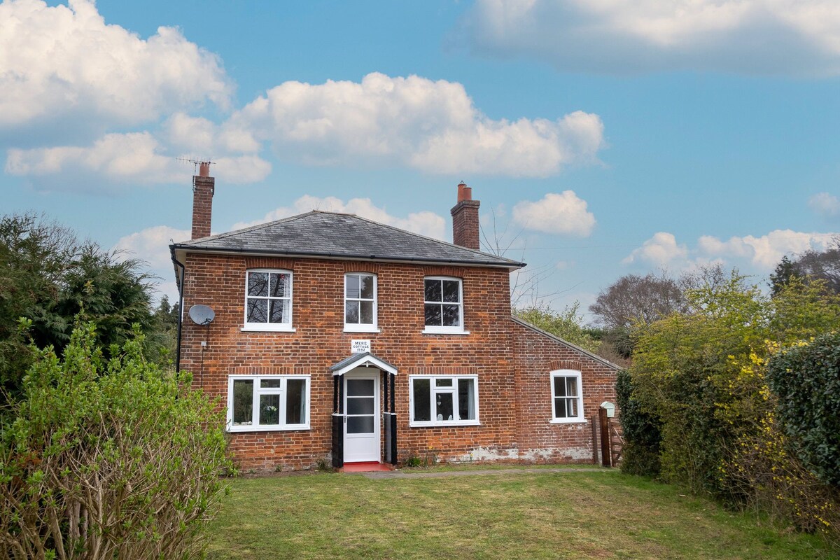 A two-story cottage is showcased amidst a green garden, featuring a brick exterior and white window accents. The front door is framed by a small porch, inviting guests into a peaceful retreat surrounded by natural greenery.