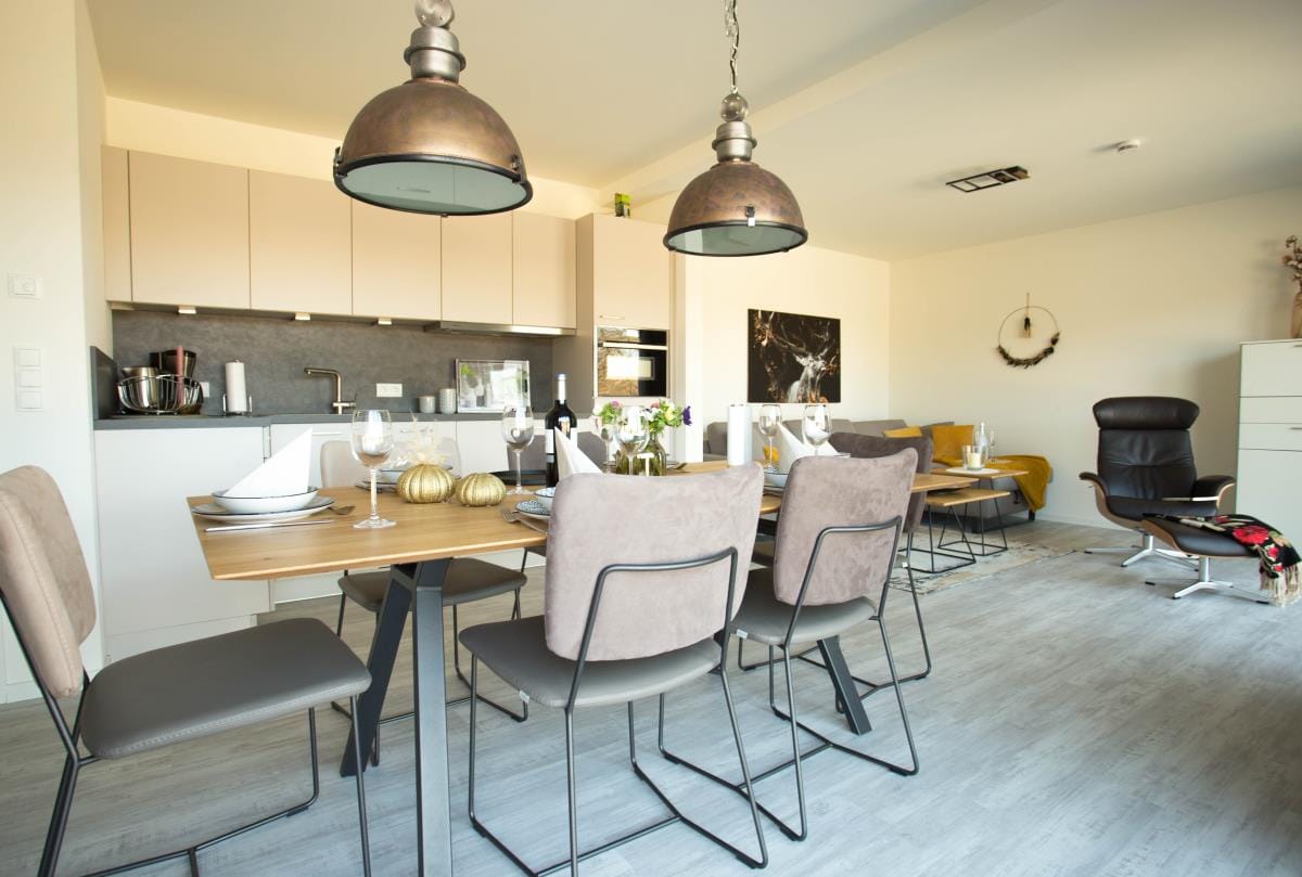 An open dining area features a wooden table accompanied by six upholstered chairs, creating a comfortable setting for meals. Pendant lights hang above, illuminating the space. A modern kitchen with light cabinetry is visible in the background, and a cozy seating area is present to one side.