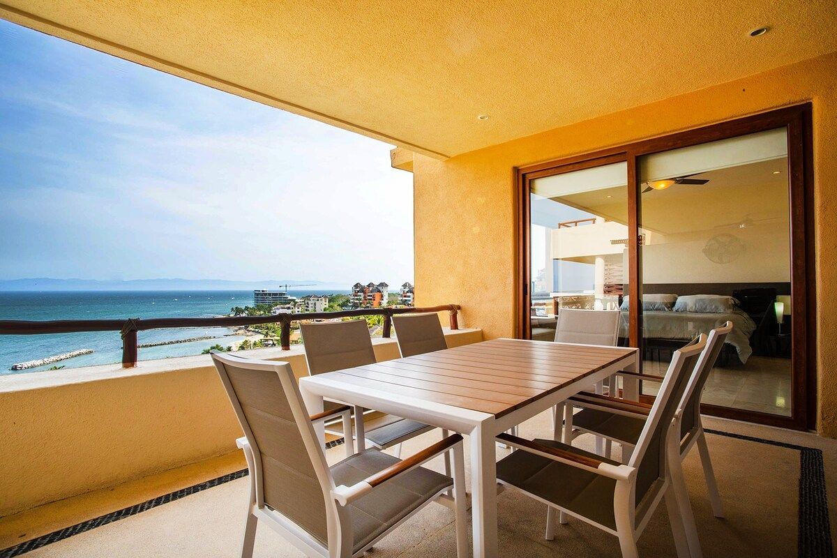 A private terrace is shown featuring a modern outdoor dining table with six chairs. The space is well-lit by natural light, offering views of the ocean and surrounding area, with sliding glass doors leading to the interior of the condominium.