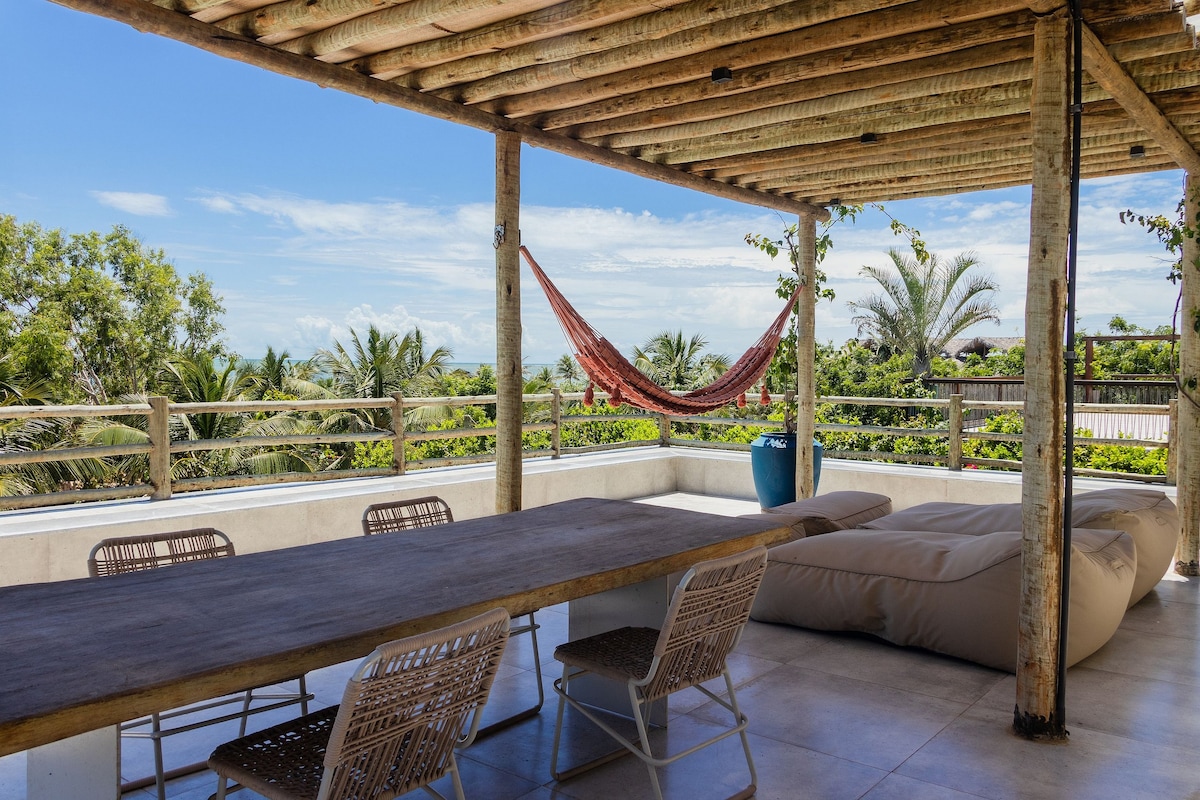 An outdoor terrace features a large wooden dining table surrounded by woven chairs. A hammock hangs nearby, while lush greenery can be seen in the background. The area is shaded by a wooden roof, providing a relaxing space to enjoy the view.