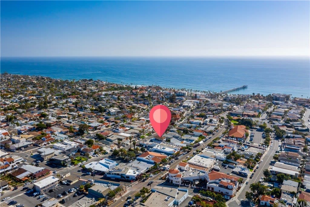 An aerial view captures a coastal area, with colorful homes lining the streets and the ocean visible in the background. A pier extends into the water, while a marker highlights the property's location in proximity to the beach.