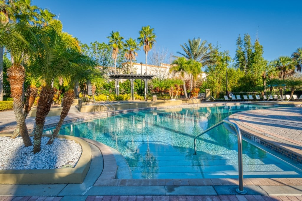 A serene pool area is surrounded by lush greenery and palm trees. The sparkling water invites relaxation, and lounge chairs are positioned along the poolside, offering ample space for sunbathing. Attention is drawn to the gentle curves of the pool, enhancing the tranquil atmosphere.