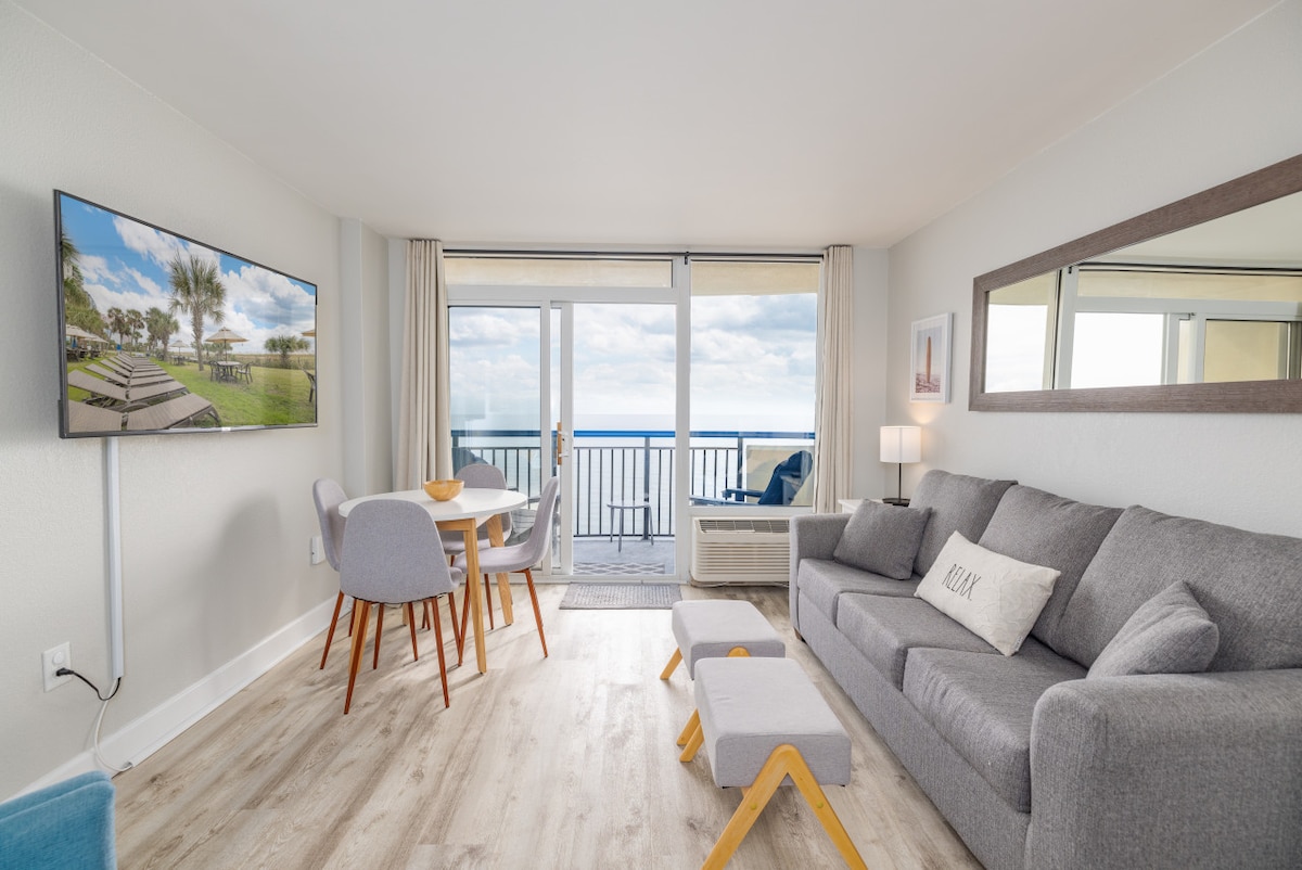 The living room features a modern gray sofa accompanied by a matching ottoman and four dining chairs around a circular table. A mounted television is positioned on the wall, and large glass doors open to a balcony, providing views of the ocean.