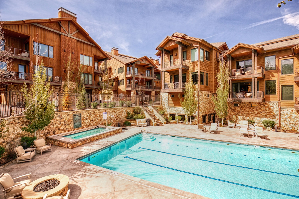 A shared outdoor area features a Junior Olympic-sized pool surrounded by a spacious deck. Comfortable lounge chairs line the poolside, and a hot tub is located nearby, all framed by well-maintained landscaping and buildings with warm wood accents.