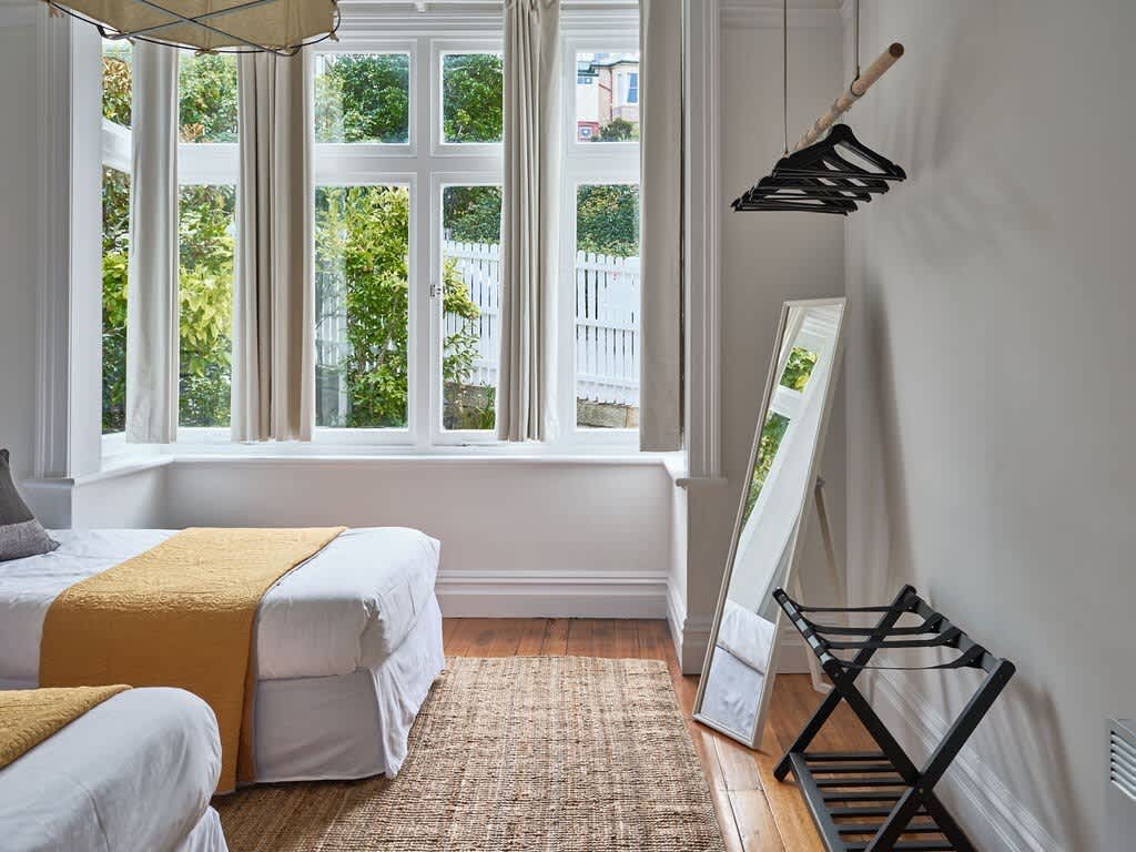 A well-lit sleeping area features two spacious beds adorned with soft yellow throws. Natural light streams in through large windows, framed by light curtains. A full-length mirror and a clothing rack are positioned beside the wall, enhancing the room's functionality.