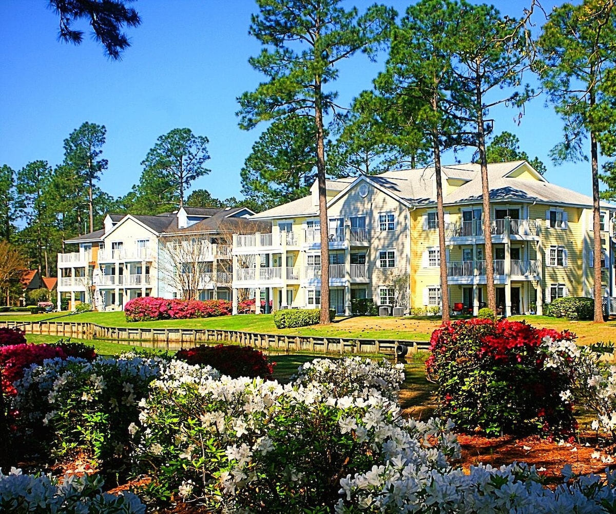 The image showcases a charming resort complex surrounded by lush greenery and colorful flowering shrubs. The two-story buildings feature balconies and wide windows, blending with the natural landscape under a clear blue sky. Tall pine trees provide shade and enhance the serene environment.