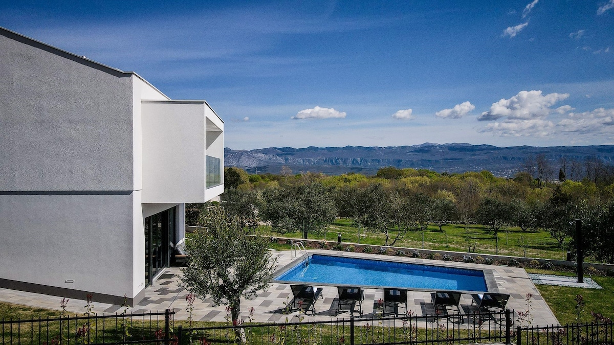 The exterior of the villa features a modern white facade, complemented by a private pool set on a spacious sun deck. Surrounding greenery adds to the serene atmosphere, while distant mountains provide a scenic backdrop under a clear blue sky.