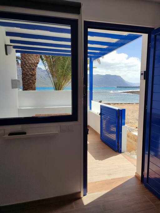 An entrance transition is visible, featuring a bright blue door open to reveal stunning beach views. A sandy area is framed by palm trees and mountains in the distance. Natural light floods the space, highlighting the inviting interior design of the room.
