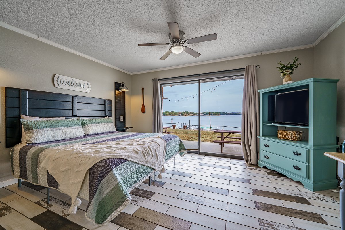 A spacious sleeping area features a large bed with a quilted cover. A ceiling fan provides circulation. Sliding glass doors open to a view of Lake Hamilton, with outdoor seating visible on the patio. A TV and a colorful dresser complete the room.