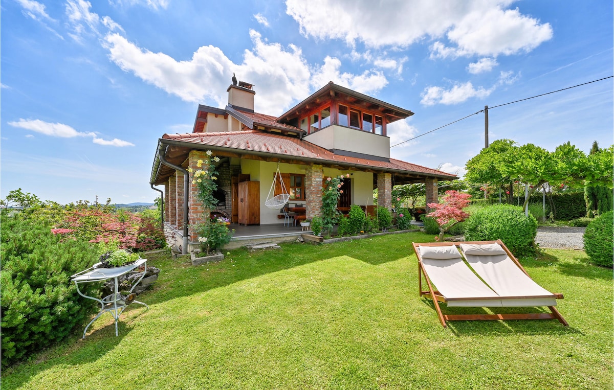 The holiday home features a welcoming facade with a sloping roof and large windows. Nestled in a landscaped garden, a terrace is highlighted with lounge chairs and a suspended hammock, surrounded by flowering plants and shrubs, offering tranquil views of the surrounding greenery.